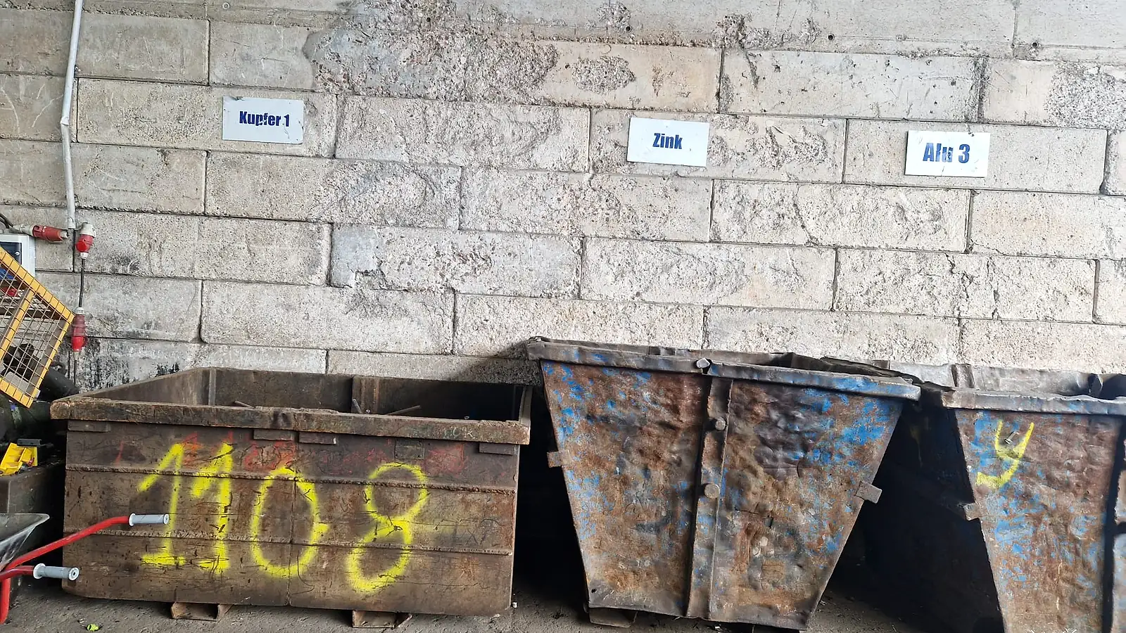 Schrottboxen vor einer Hallenwand mit den Sortenschildern Kupfer 1, Zink und Alu 3